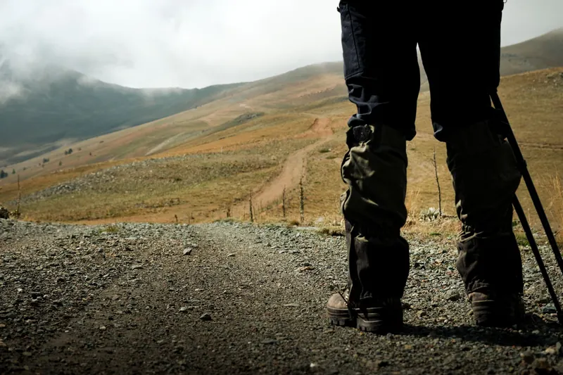 A hiker stands on a rocky mountain trail with walking poles, facing vast hilly landscapes