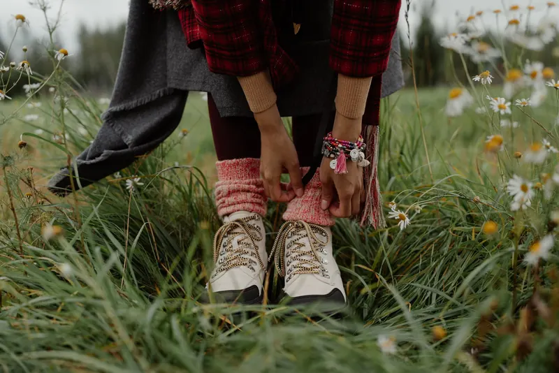 A woman in a plaid jacket ties her hiking boots surrounded by wild daisies in a meadow