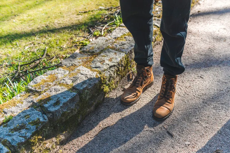 A person stands on a sunlit path wearing stylish leather boots and dark pants